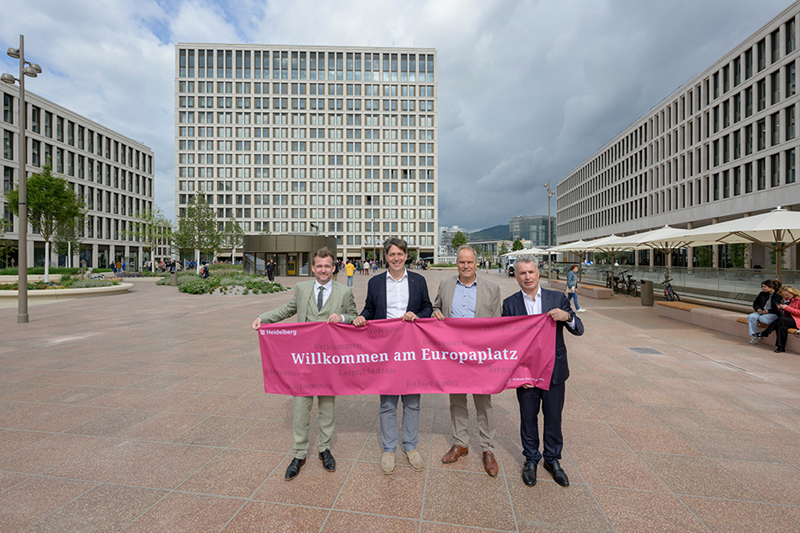 Vier Männer in Anzug halten ein heidelbeerfarbenes Banner mit der Aufschrift "Willkommen am Europaplatz"